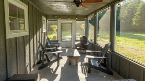 Área externa coberta com ventilador de teto, cadeiras de madeira, mesa e grandes janelas que oferecem vista para um campo gramado.