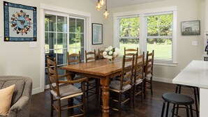 Uma sala de jantar com mesa de madeira, seis cadeiras, um vaso com flores e uma janela grande com vista para o verde.