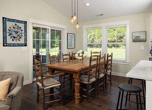 Un coin repas avec une table en bois, six chaises, un vase de fleurs et une grande fenêtre donnant sur un paysage verdoyant.