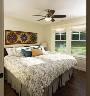 Une chambre à coucher comprenant un lit, un ventilateur de plafond, une fenêtre donnant sur un paysage verdoyant et une tapisserie décorative au mur.