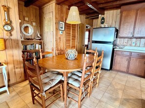 A wooden dining table with chairs, a hanging light fixture, a bookshelf, and a refrigerator.