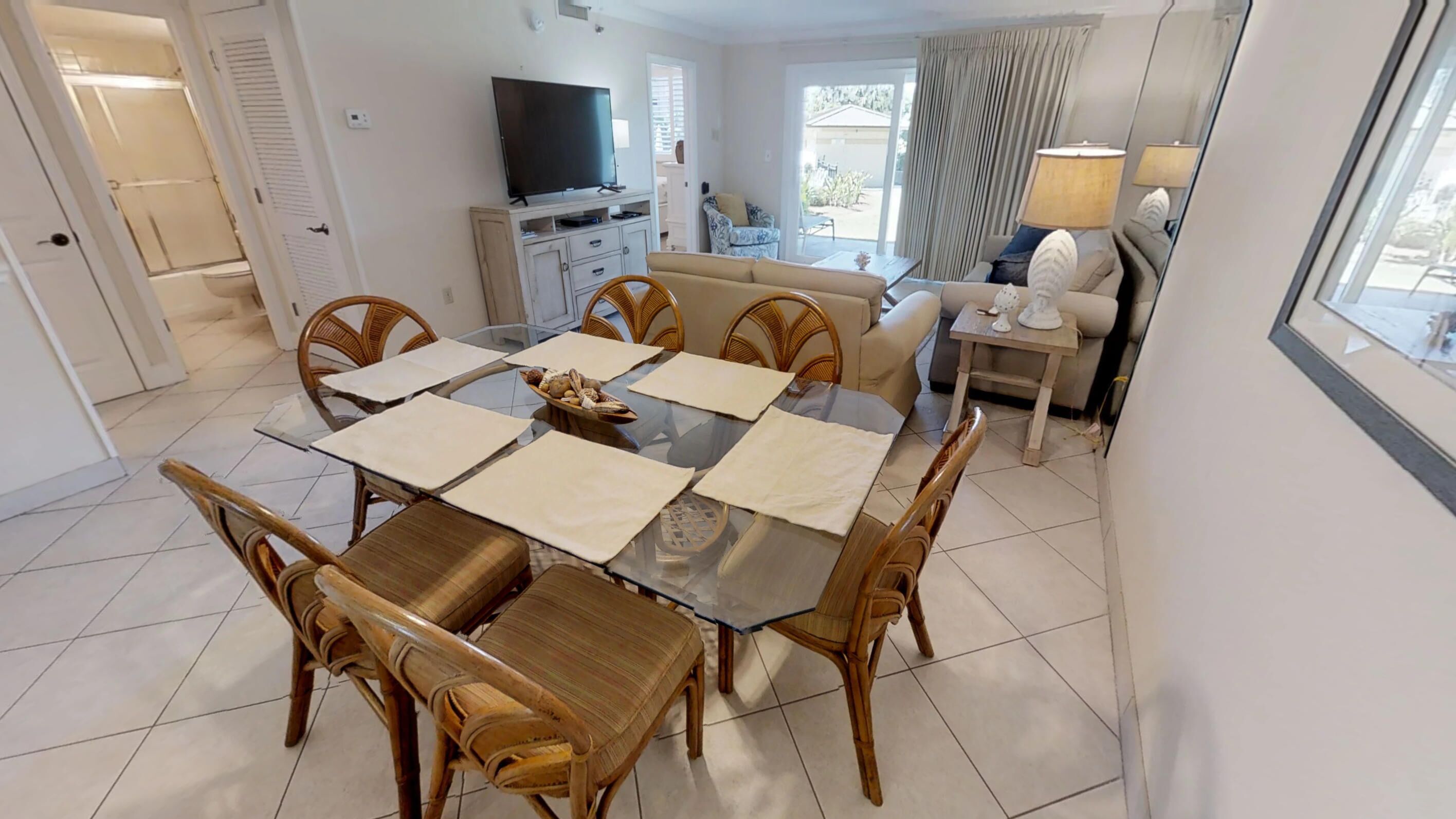 A dining area with a glass table, wooden chairs, and a television in the background.