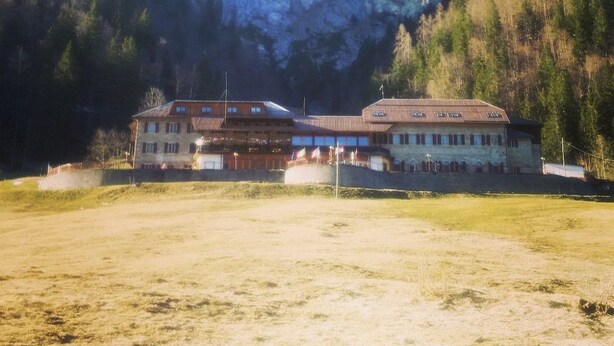 Un bâtiment doté de nombreuses fenêtres et d’un toit plat, entouré d’une zone verdoyante et avec des montagnes en arrière-plan.