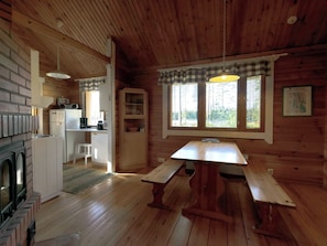 A cabin interior with a wooden floor, a dining table with benches, a kitchen area with appliances, and a window with blinds.