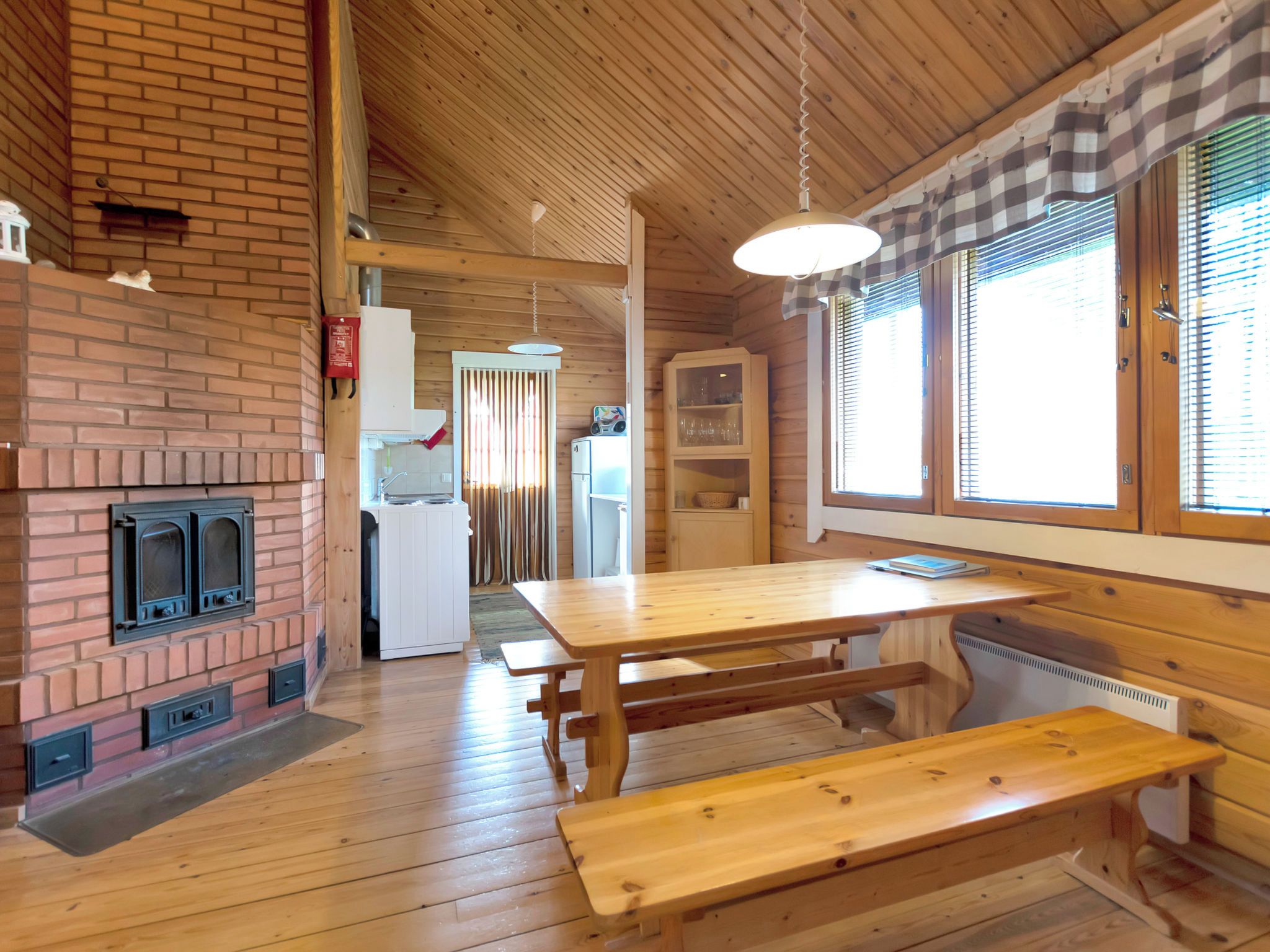En inredning med träpanel, en tegelbeklädd öppen spis, ett matbord med bänkar och ett köksområde.