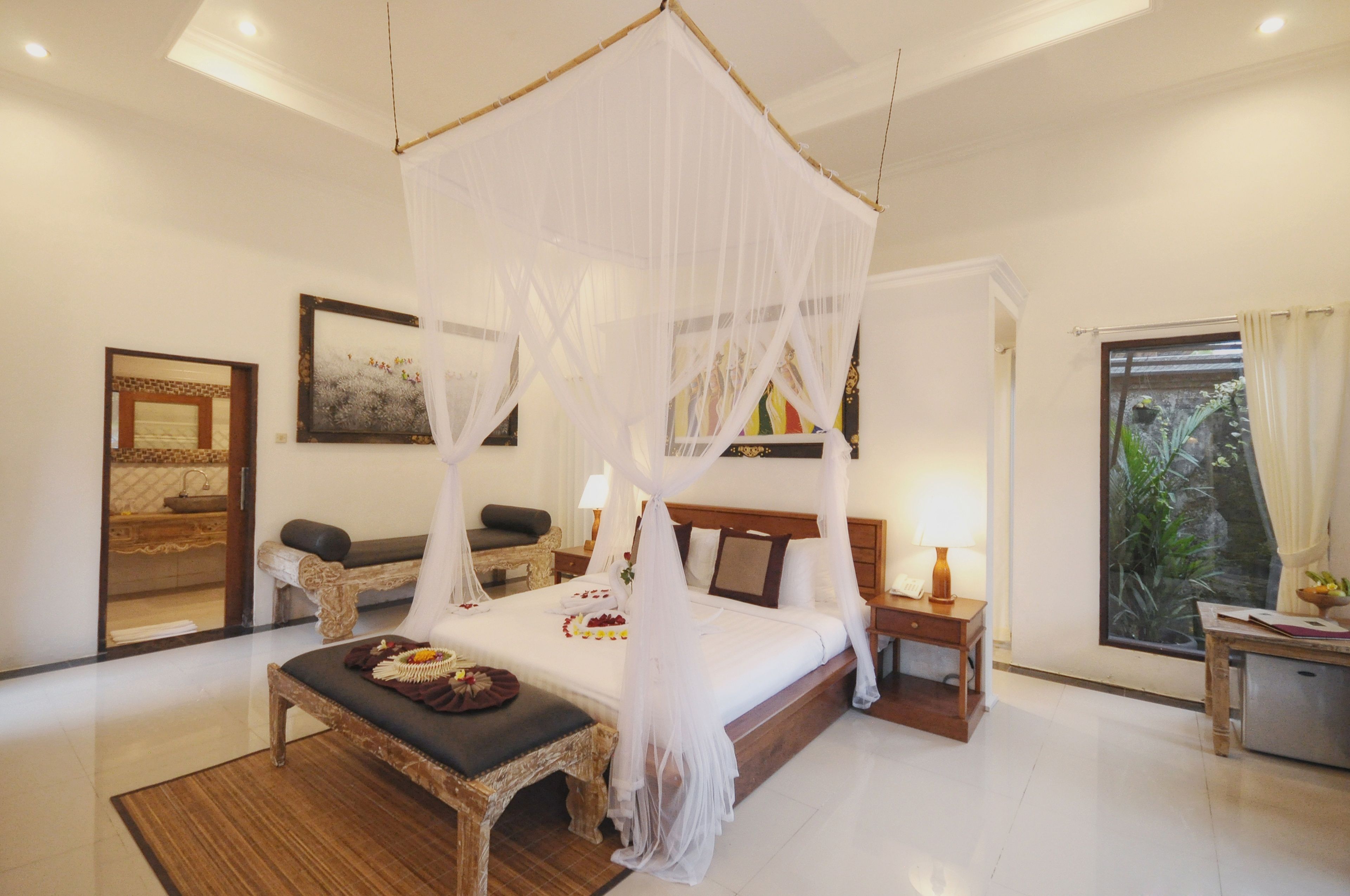A bedroom with a four-poster bed, a sitting area with a bench, a bedside table with a lamp, and a window with curtains.