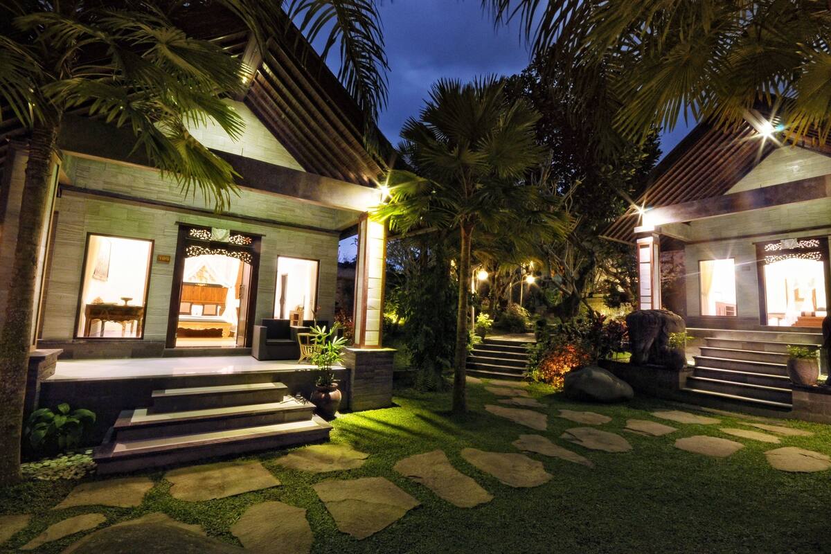 A modern house with a well-lit entrance, stone steps, and a garden with palm trees and a stone pathway.
