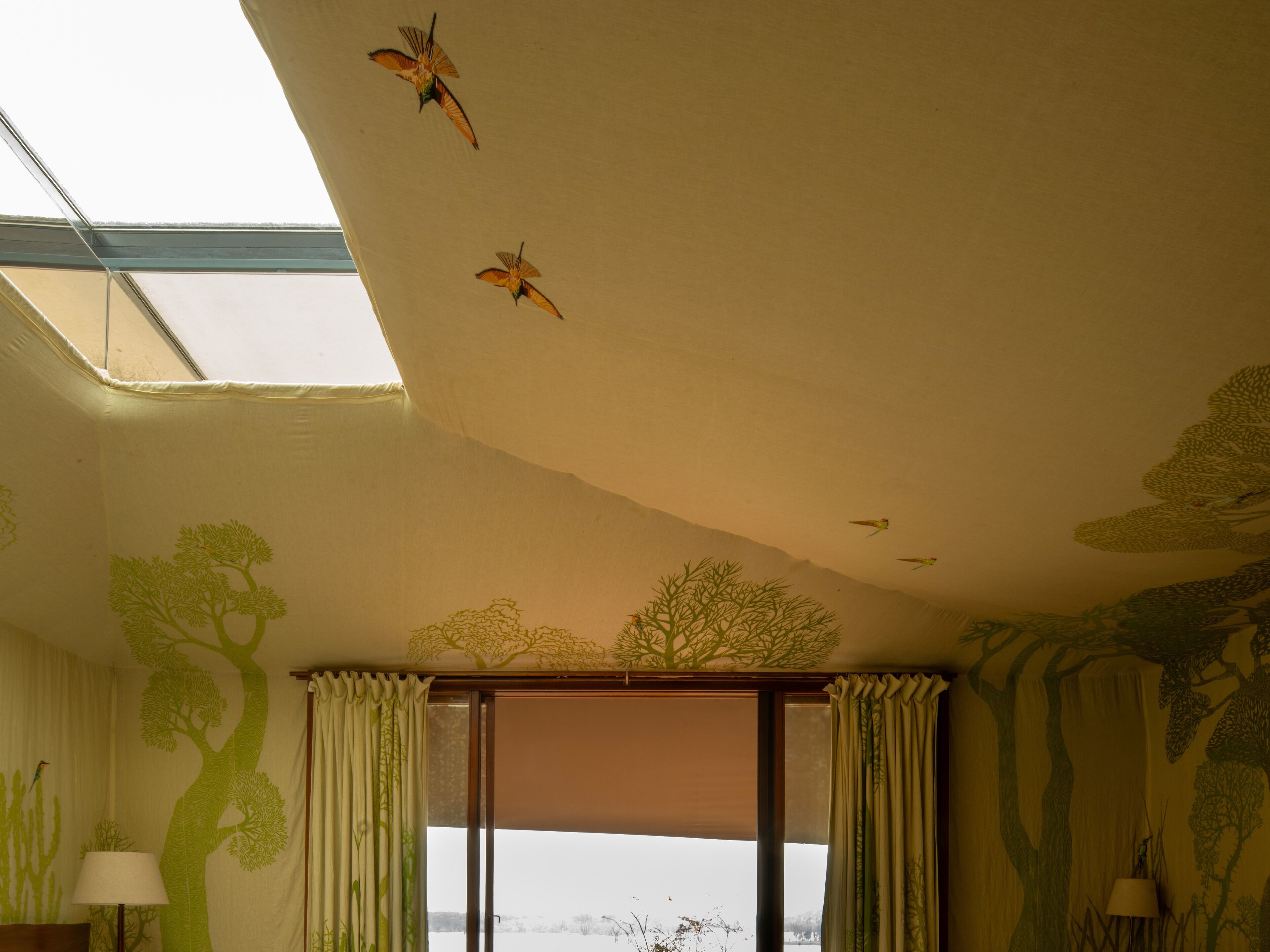 Quarto com claraboia, vista para o exterior e um mural de árvore na parede.