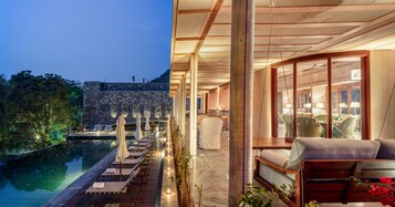 Un área de piscina exterior lujosa con sillones de descanso y sombrillas, un muelle de madera y vistas a un muro de piedra y árboles.