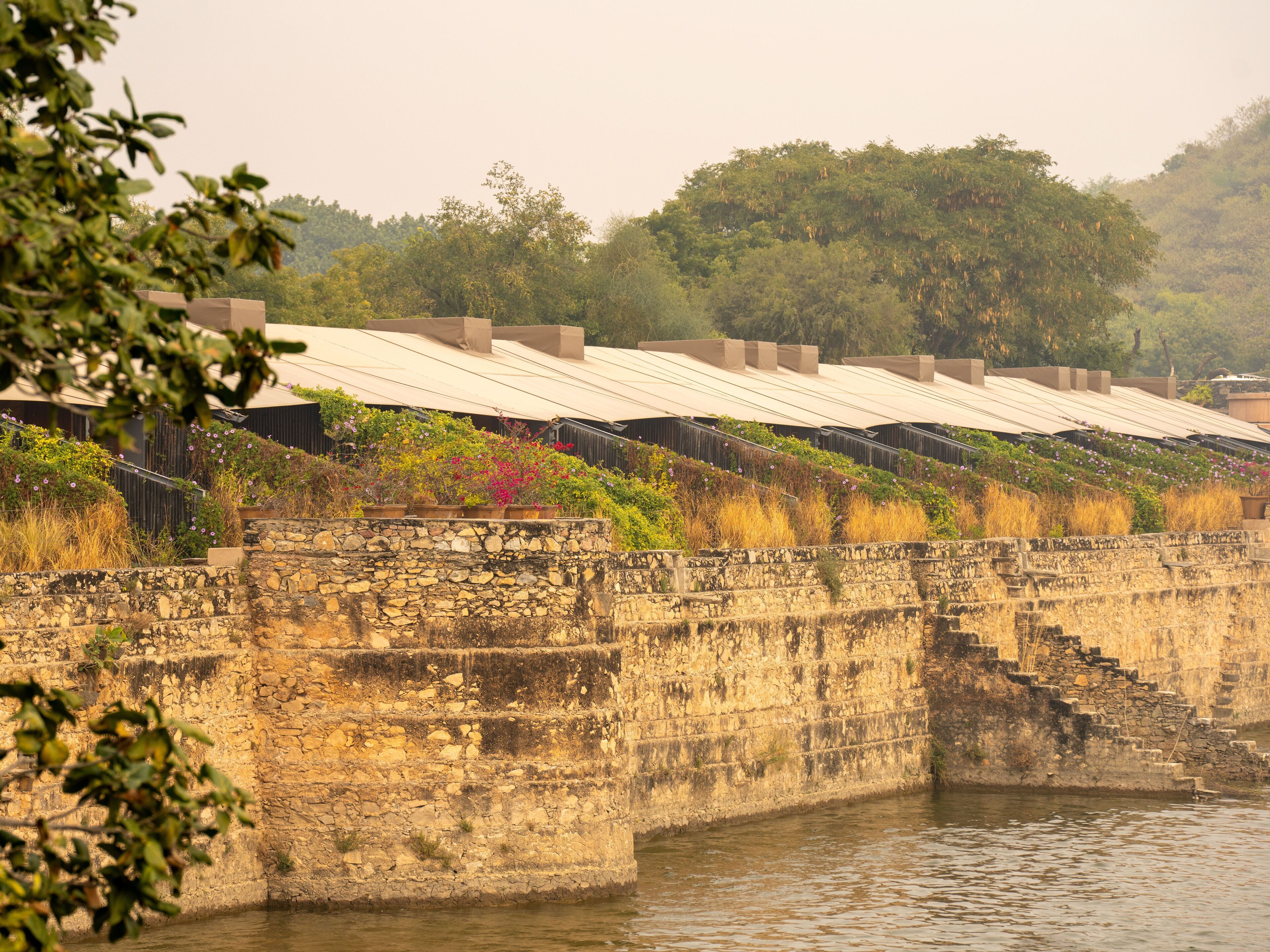 A stone retaining wall with a water body in front, a staircase, and a row of buildings with flat roofs.