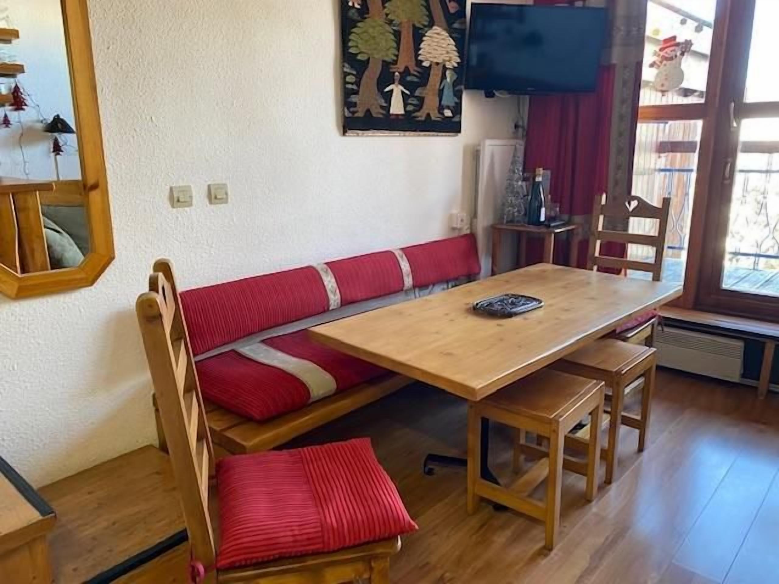 Una habitación con una mesa de madera, un banco tapizado en rojo y un televisor montado en la pared.