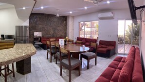 Una cocina moderna con encimeras de granito, un refrigerador empotrado y una cafetera. Hay un comedor con una mesa de madera y sillas tapizadas en rojo. Una pared de piedra separa la cocina del comedor.