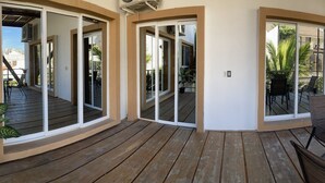 Una terraza de madera con puerta de vidrio que da a un balcón, una mesa redonda con sillas y una planta en maceta.