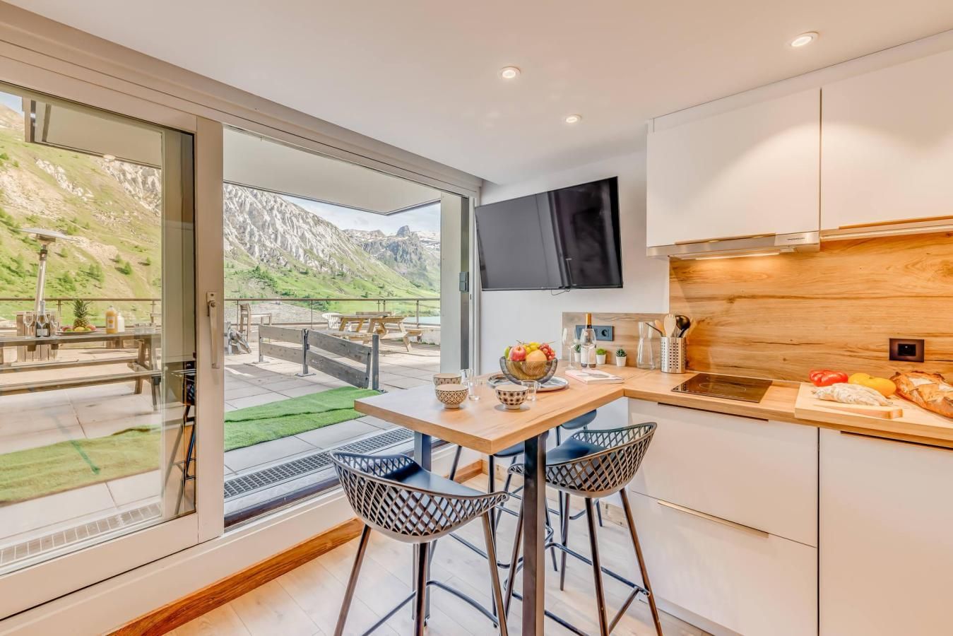 A modern kitchen with a large window overlooking a mountainous landscape, a dining area with two chairs, and a wooden countertop with kitchen utensils.