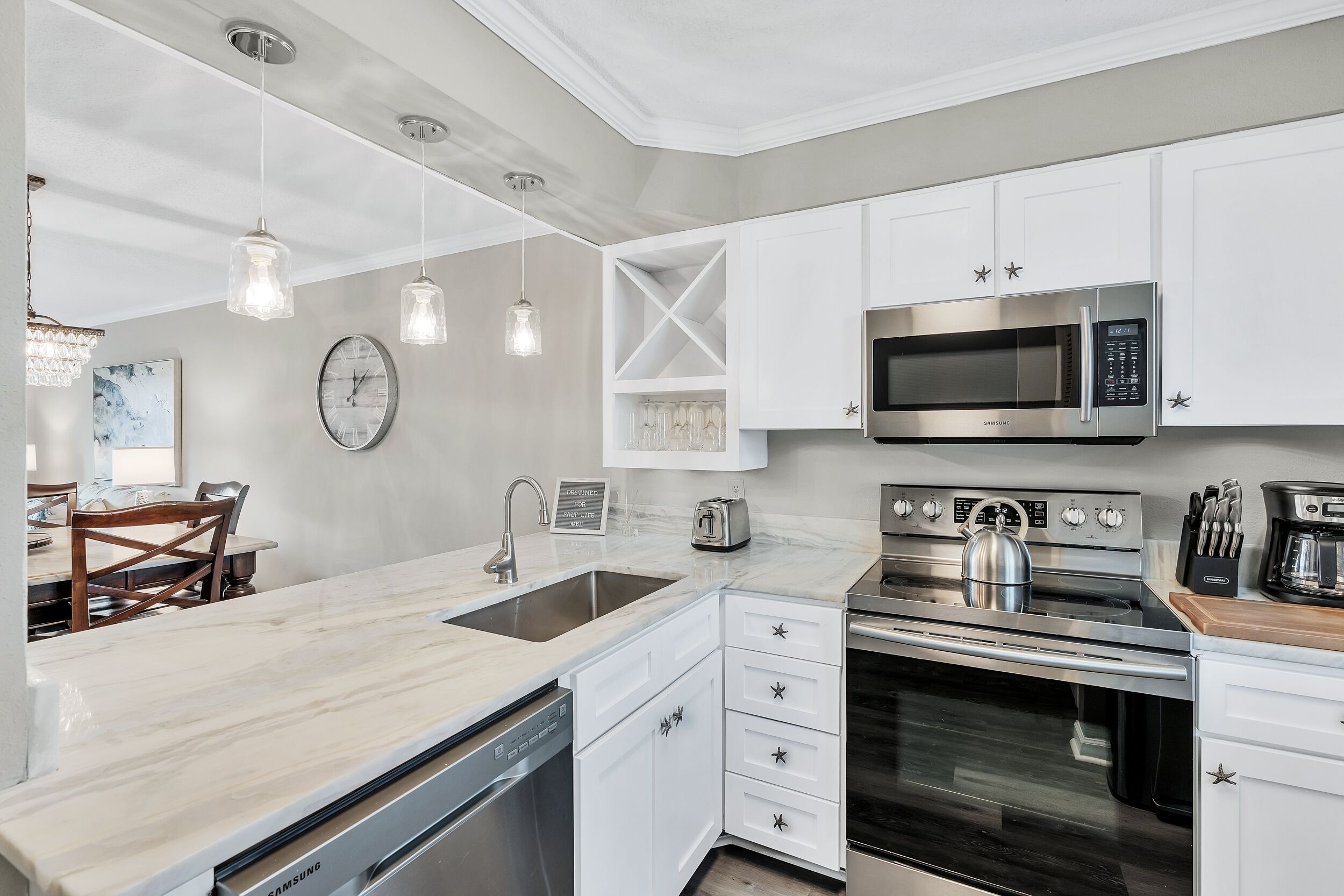 A modern kitchen with white cabinetry, a stainless steel microwave, an oven, and a dishwasher. There are pendant lights, a clock, and a starfish decoration on the cabinets.