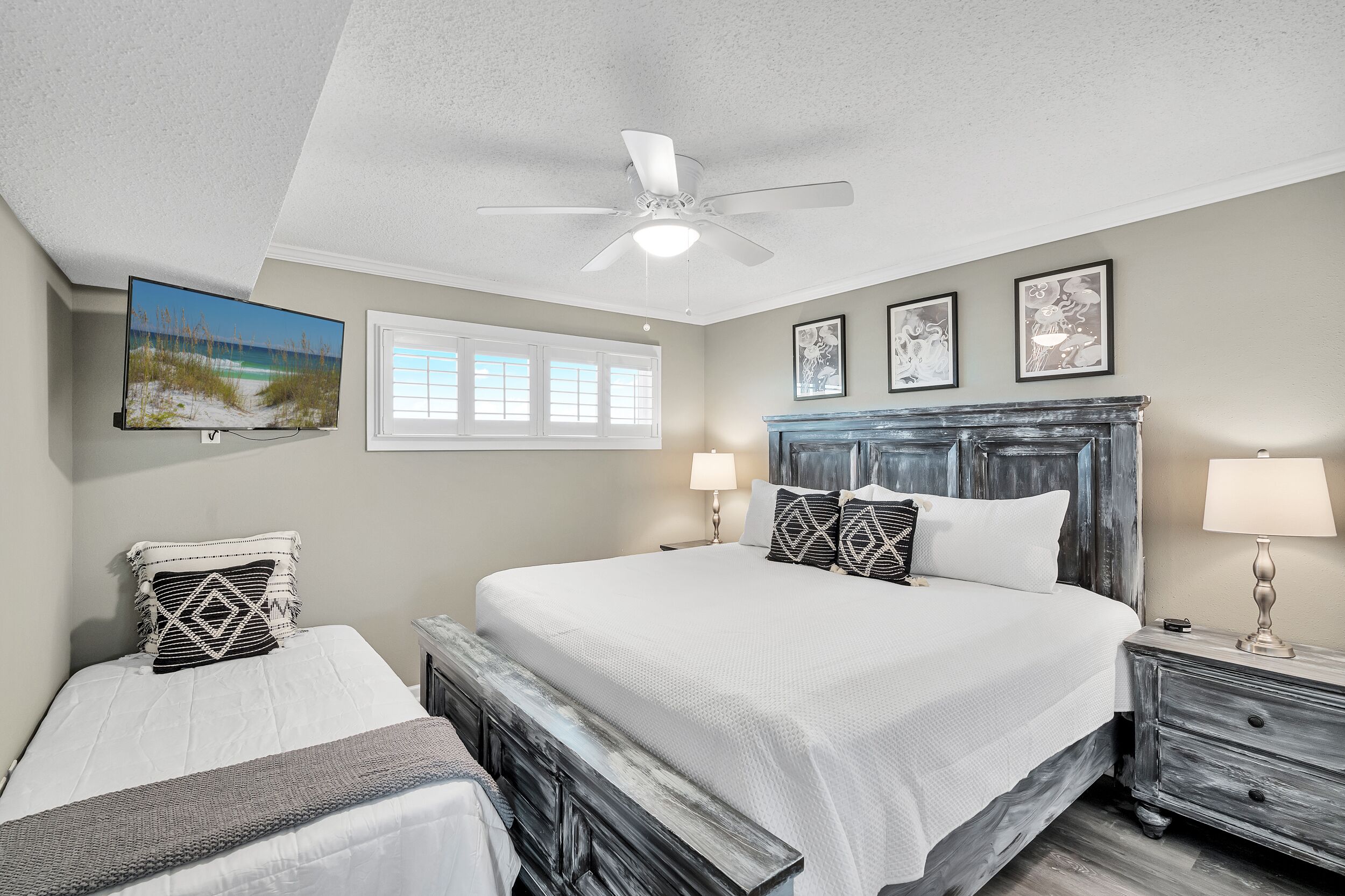 A bedroom with a large bed, a smaller bed, a ceiling fan, a TV, and artwork on the walls.