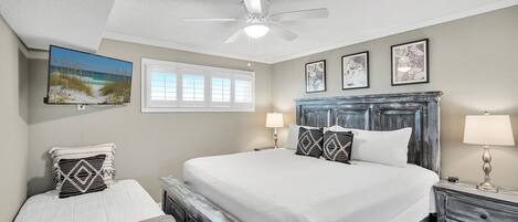 A bedroom with a large bed, a smaller bed, a ceiling fan, a TV, and artwork on the walls.