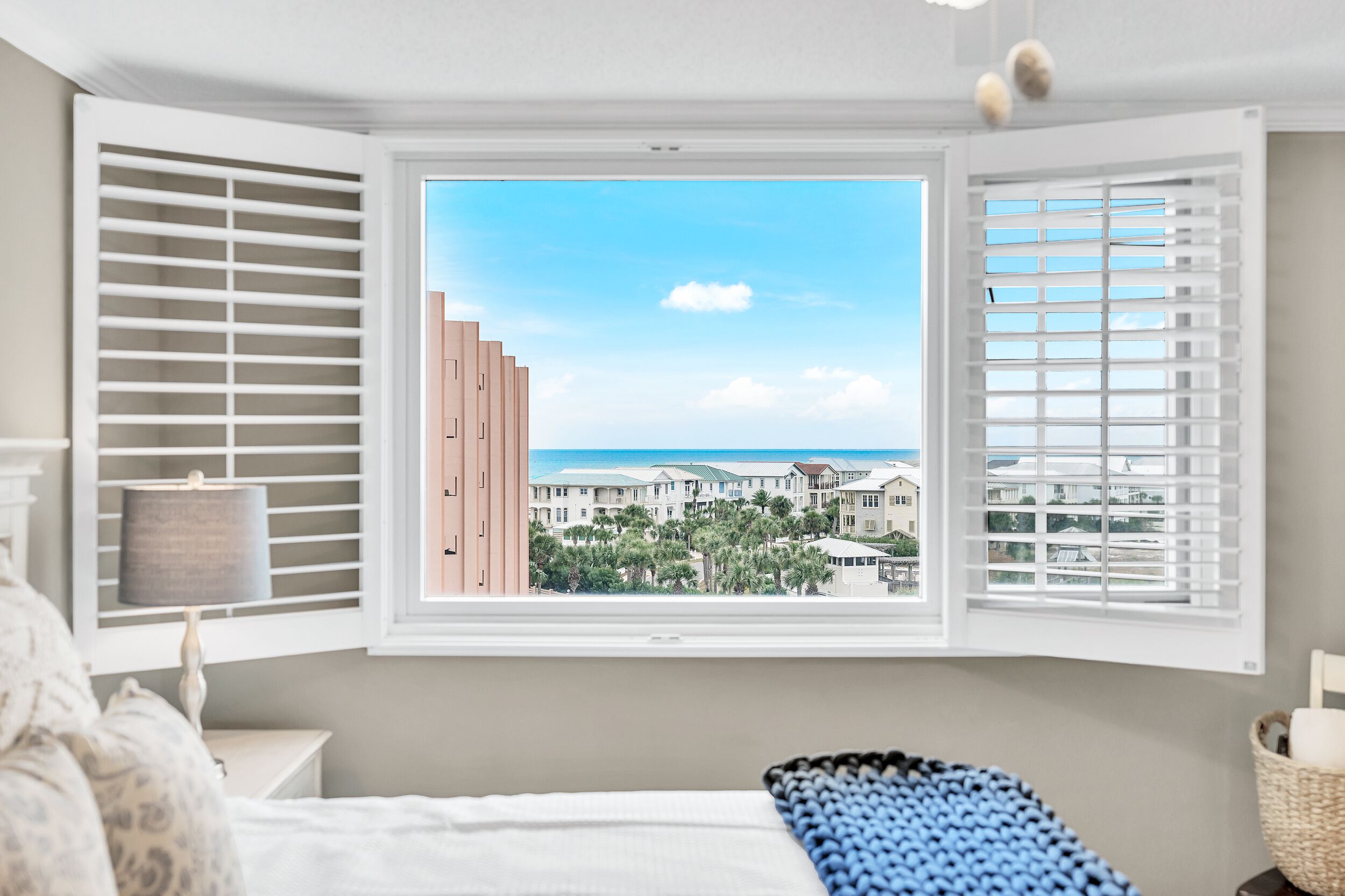 A bedroom with a large window, white blinds, a bed, a bedside lamp, and a view of a building and trees.
