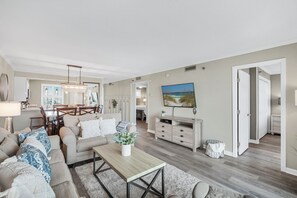 Ein geräumiges Wohnzimmer mit einer Sofagarnitur, einem Couchtisch und einem Fernseher. Im Hintergrund ist eine Essecke mit Tisch und Stühlen zu sehen.