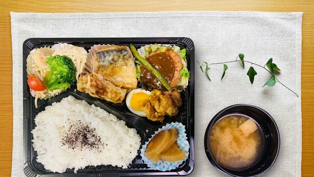 Una caja bento con arroz, pescado, huevo y verduras sobre una mesa, junto a una servilleta y un tazón pequeño de sopa.