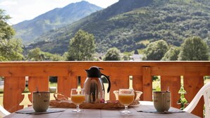 A table with a coffee pot, two glasses of orange juice, and two cups on a wooden deck with a mountain view in the background. - Wifi, view mountain, swimming pool, sauna, hammam, terrace, balcony, parking, tv, ski locker, 38m² (Luz-Saint-Sauveur)