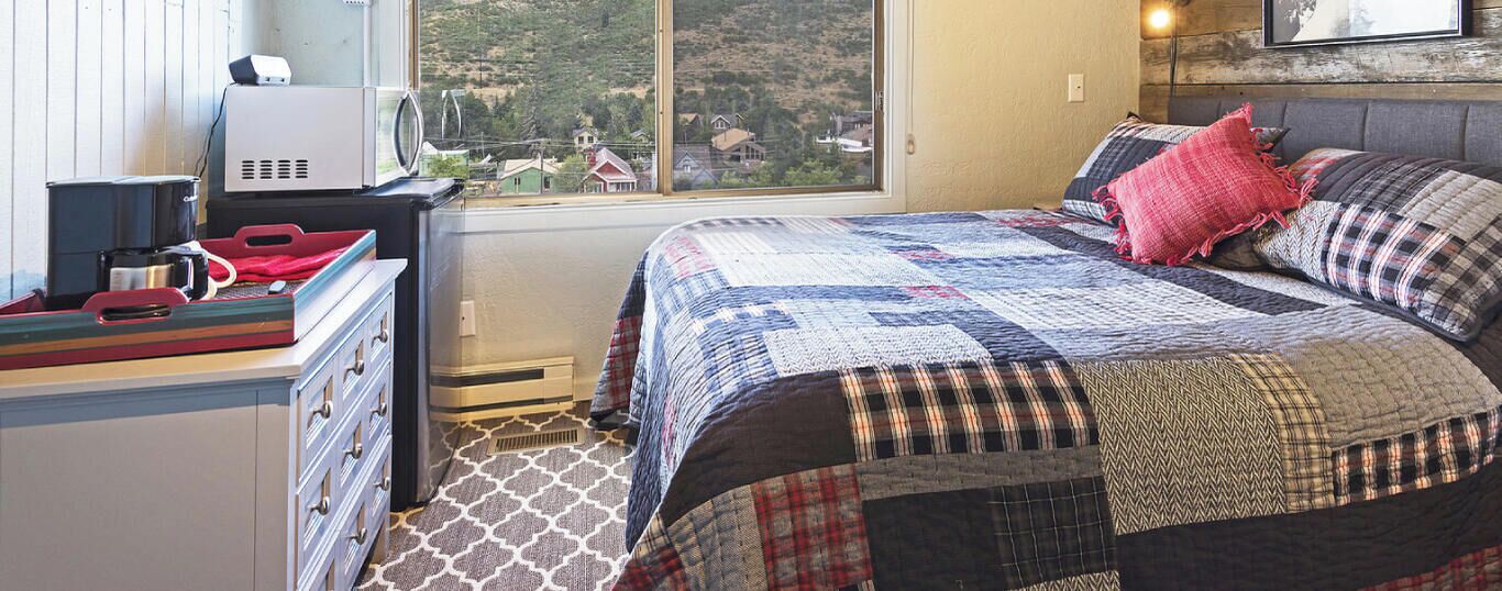 A bedroom with a large window, a bed with a quilt, a desk with a computer, and a kitchen area.