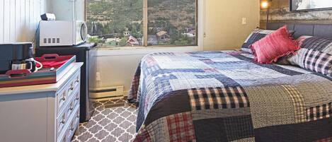 A bedroom with a large window, a bed with a quilt, a desk with a computer, and a kitchen area.
