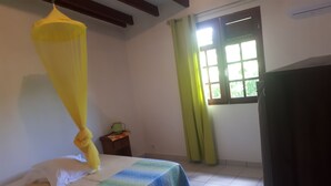 A room with a bed, a window with curtains, a small table with a plant, and a yellow hanging canopy. - Charming cozy house type T3---------------------- (sainte-anne en Guadeloupe)