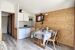 Een kleine keuken met witte kastjes, een magnetron en een spoelbak. Een eettafel met stoelen staat tegen een houten wand.