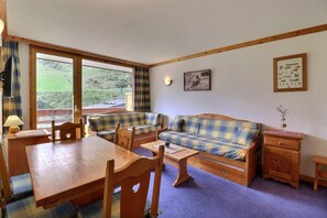 A cozy room with a wooden dining table, two plaid sofas, a small wooden cabinet, and a window with a view of greenery. - 10m from ski slopes, 6th floor, balcony, 40m², Méribel-Mottaret (Méribel - Les Allues)