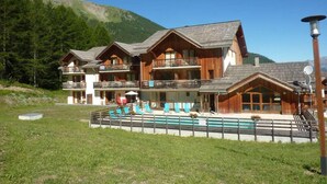 A chalet-style hotel with multiple balconies, a pool area, and a mountainous backdrop.