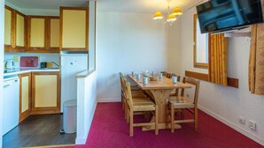 Une cuisine de taille réduite, dotée de meubles en bois, d’un réfrigérateur et d’un évier. On y trouve également un coin repas avec une table et des chaises en bois, ainsi qu’un téléviseur fixé au mur.