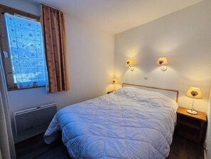 A bedroom with a bed, two bedside lamps, a window with curtains, and a wall-mounted heater.