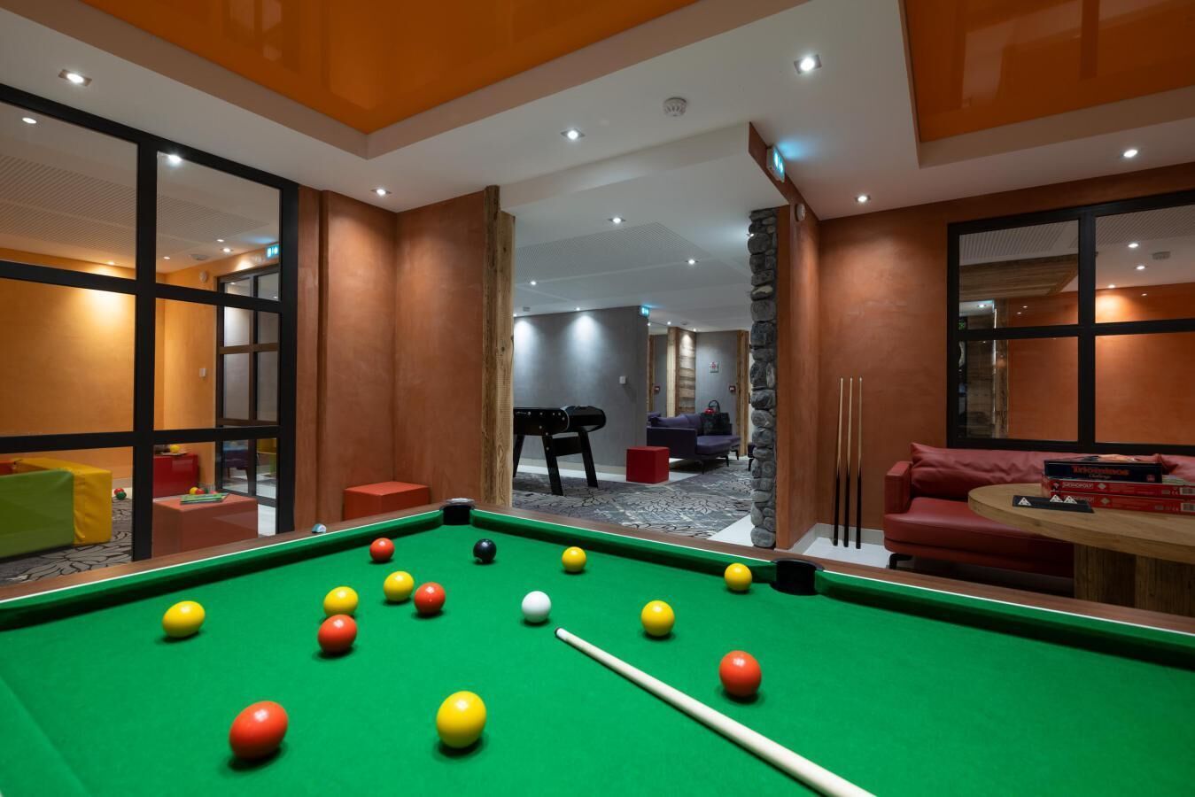 Une table de billard avec des billes, un canapé et une table avec des livres dans une pièce moderne aux murs en bois et aux grandes fenêtres.