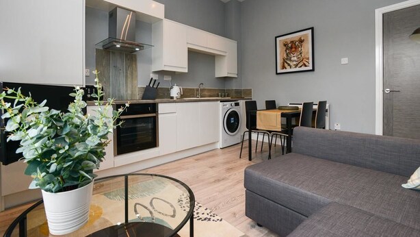 A modern kitchen with white cabinets, a built-in oven, and a washing machine. There is a grey sofa and a dining table with chairs in the living area. - Arena Apartments (Nottingham)