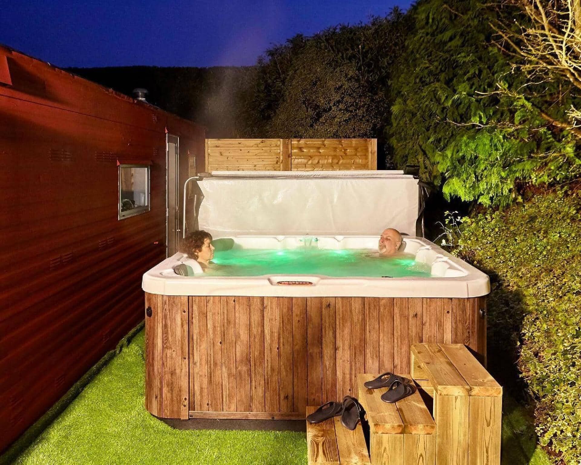 A wooden hot tub with two people inside, surrounded by a wooden deck and greenery.