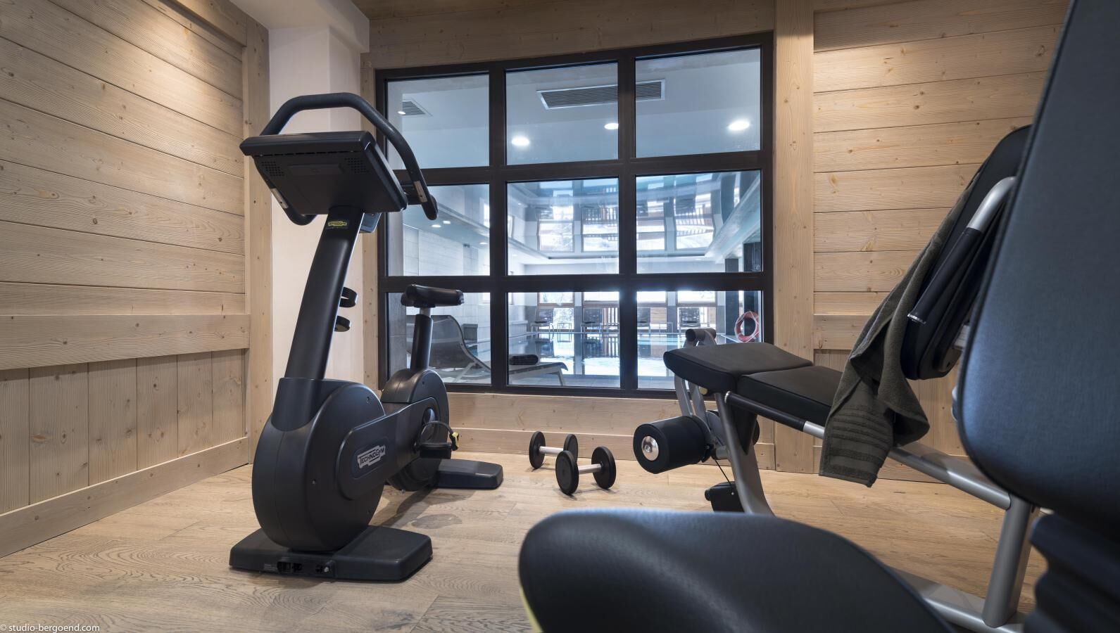 A gym with exercise equipment, a bench, and a window with a view of a building outside.