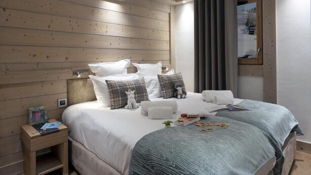 Une chambre à coucher avec une tête de lit en bois, un lit recouvert de draps blancs et agrémenté de plusieurs oreillers, une table de chevet avec un livre et une petite lampe, et une fenêtre avec des rideaux.