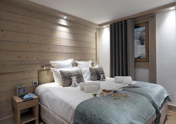 A bedroom with a wooden headboard, a bed with white bedding and multiple pillows, a bedside table with a book and a small lamp, and a window with curtains.
