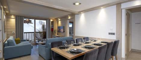 Un espace repas moderne avec une longue table dressée pour six personnes, un canapé et un balcon offrant une vue sur un paysage enneigé.