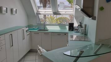 Une cuisine moderne avec des meubles blancs, un plan de travail en verre, un lave-vaisselle encastré et une fenêtre avec des stores.