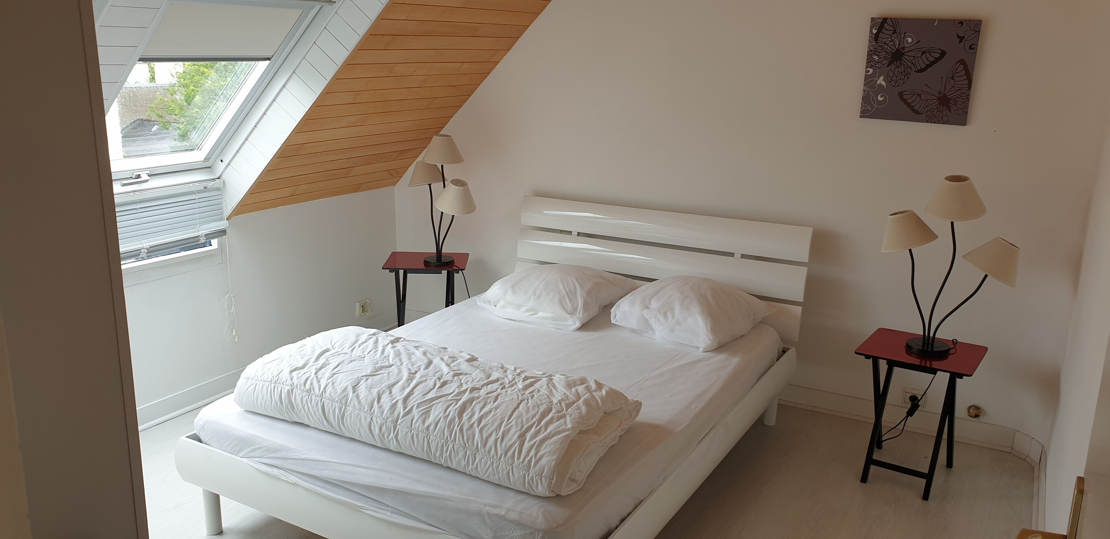 Un dormitorio con techo inclinado, una cama con ropa de cama blanca, dos mesitas de noche con lámparas y una obra de arte con temática de mariposas en la pared.