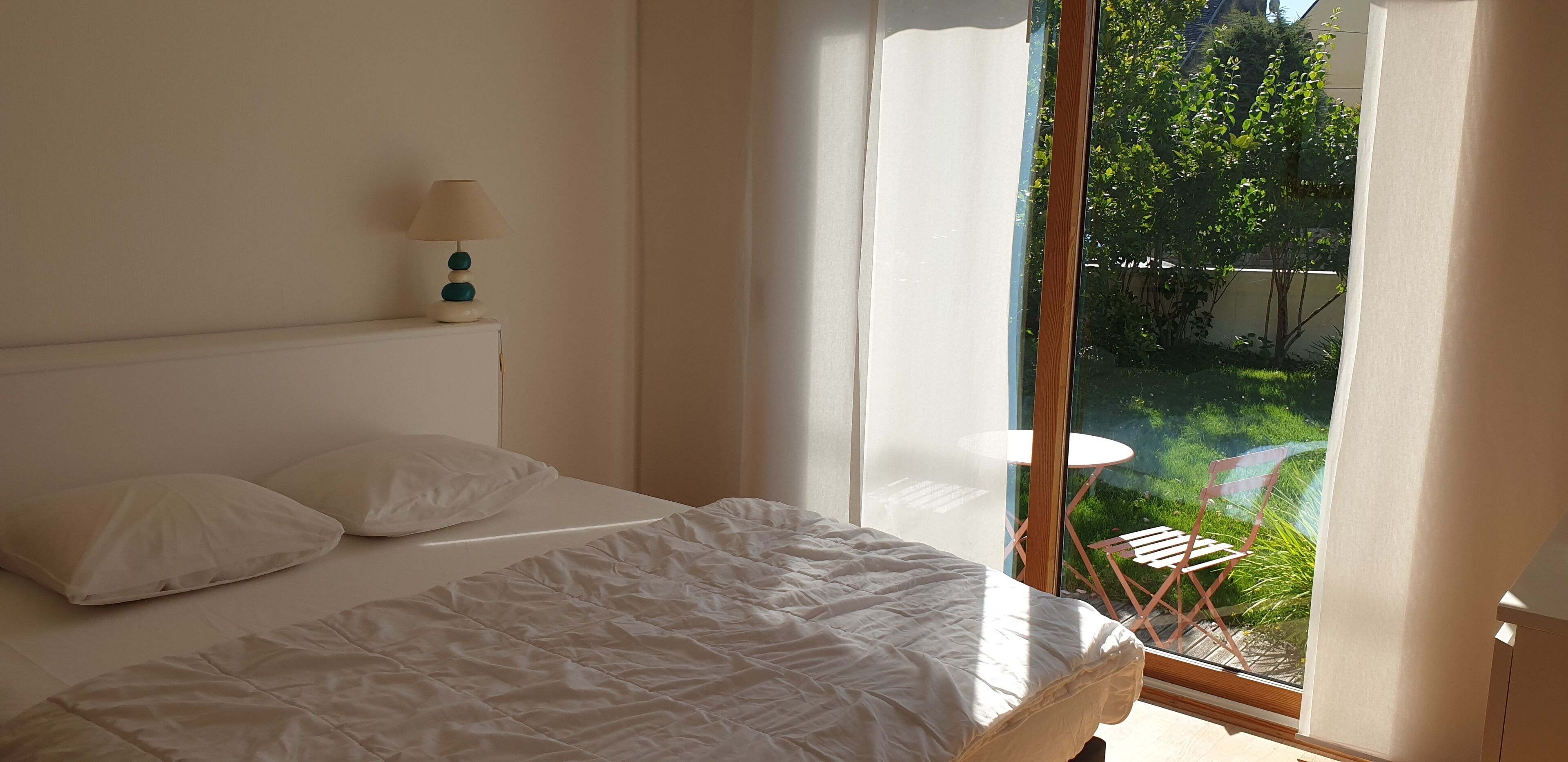 Een slaapkamer met een bed, een nachtkastje, een lamp en uitzicht op een tuin door een schuifdeur.