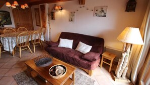 A cozy living room with a wooden dining table, chairs, a sofa with cushions, a coffee table with decorative items, and a lamp.