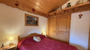 A bedroom with a wooden ceiling, a bed with a heart-shaped pillow, a nightstand with a lamp, and a wooden wardrobe.
