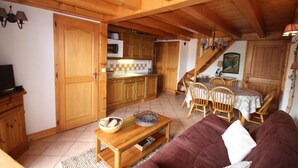 A cozy living room with a wooden ceiling, a dining area with a table and chairs, and a kitchen with wooden cabinets and a microwave.
