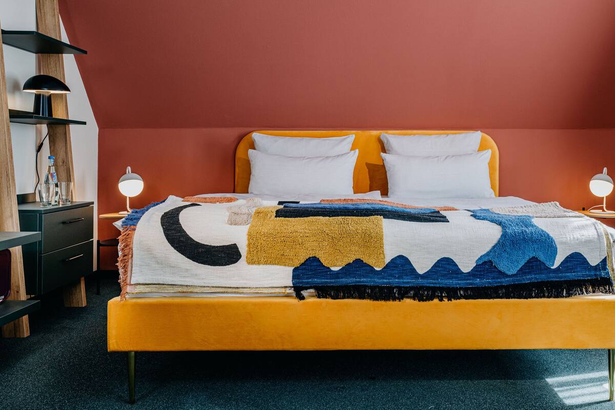 A bed with a colorful blanket, a red ottoman, and a wooden nightstand with a lamp.