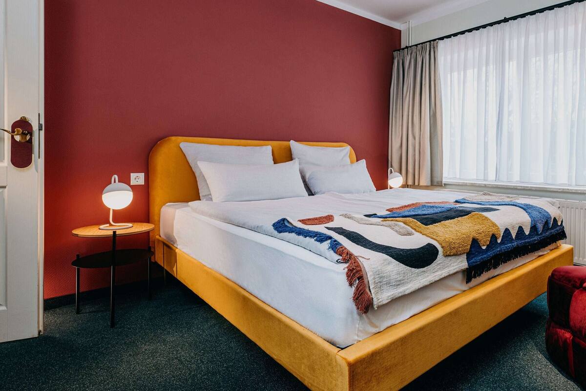 A bed with a patterned blanket, a bedside lamp, a red ottoman, and a yellow chair in a room with red walls and a window with curtains.