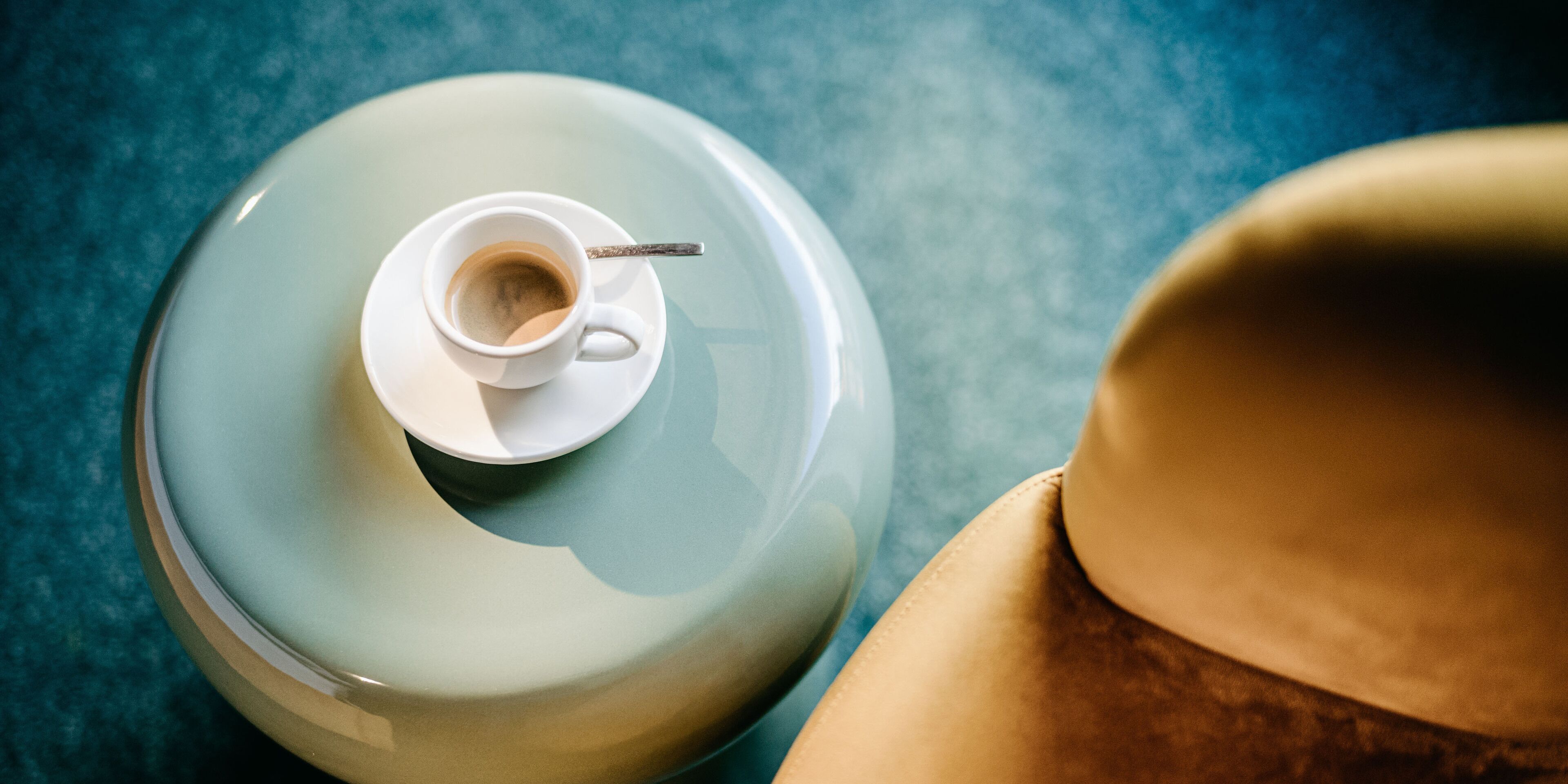 Uma chávena de café branca sobre um pires, uma colher e um chapéu bege numa superfície azul.