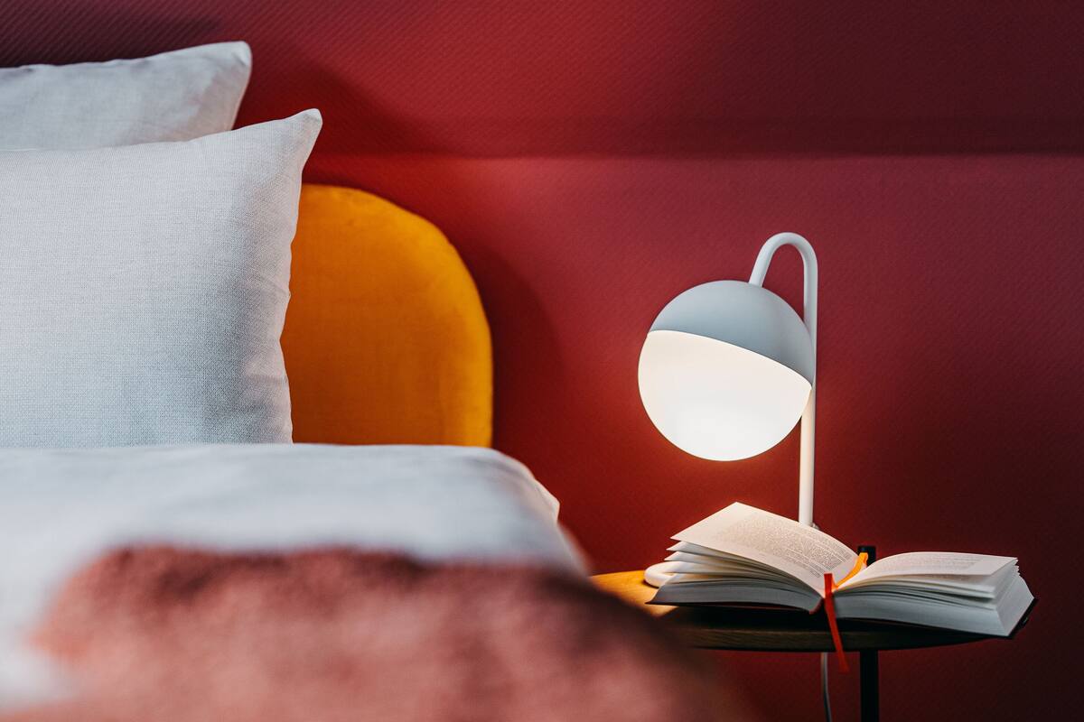 A bed with white and orange pillows, a bedside table with a lamp, and an open book.